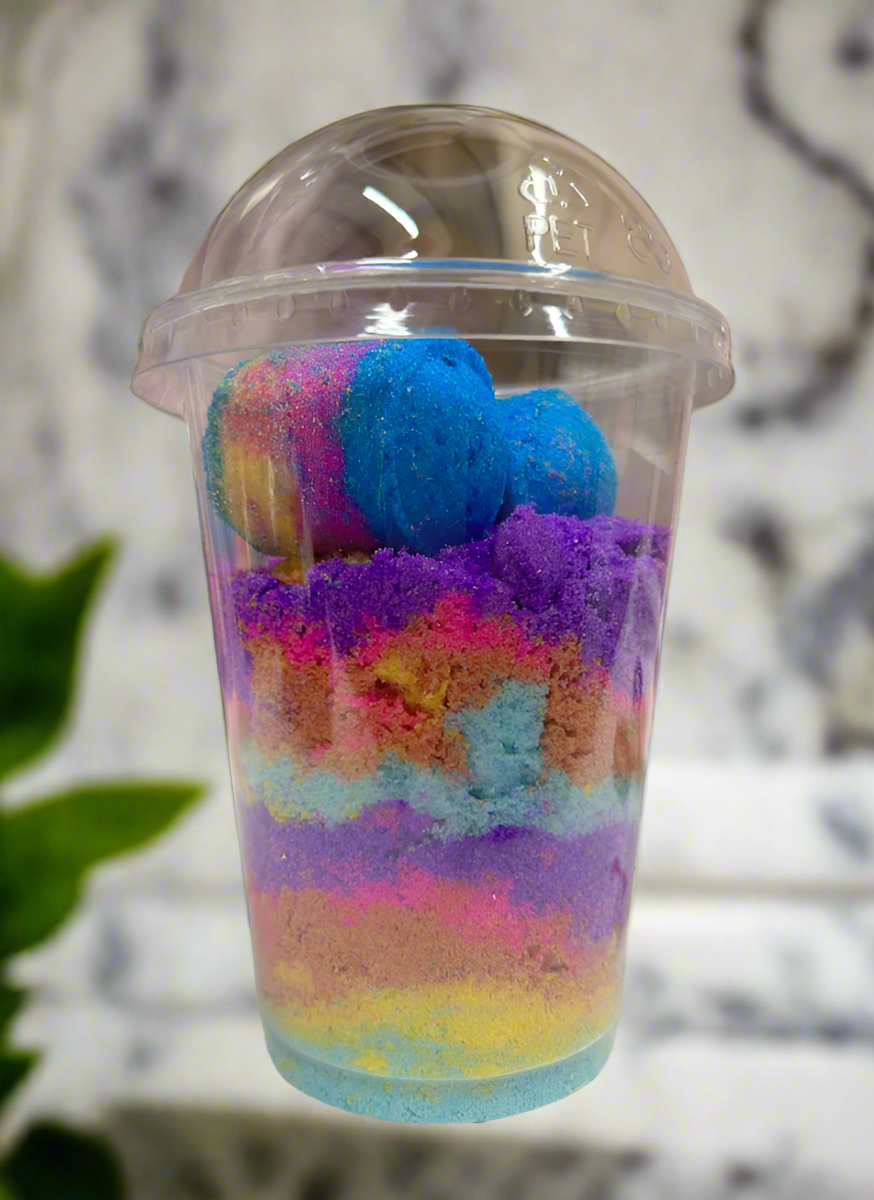 Colorful layered sand in a clear plastic cup with a lid, set against a neutral background.